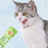 Gato doméstico gris y blanco lamiéndose los labios frente a una cuchara exprimidora verde claro que contiene una porción de snack crema